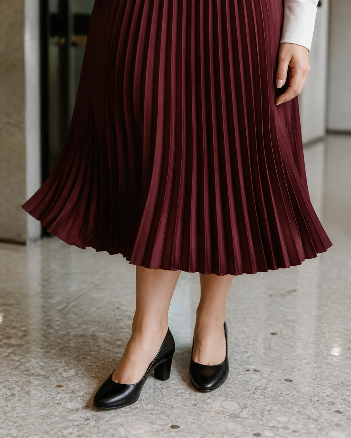 A close crop showing the hem of a burgundy pleated midi skirt with accordion pleats catching the light, and black block-heel pumps on a pale terrazzo floor