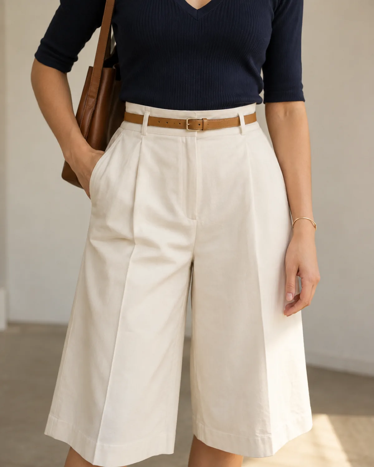 Close crop of a petite woman in a navy ribbed V-neck knit tucked into ivory pleated wide-leg culottes with a thin tan belt, a brown leather bag on her shoulder