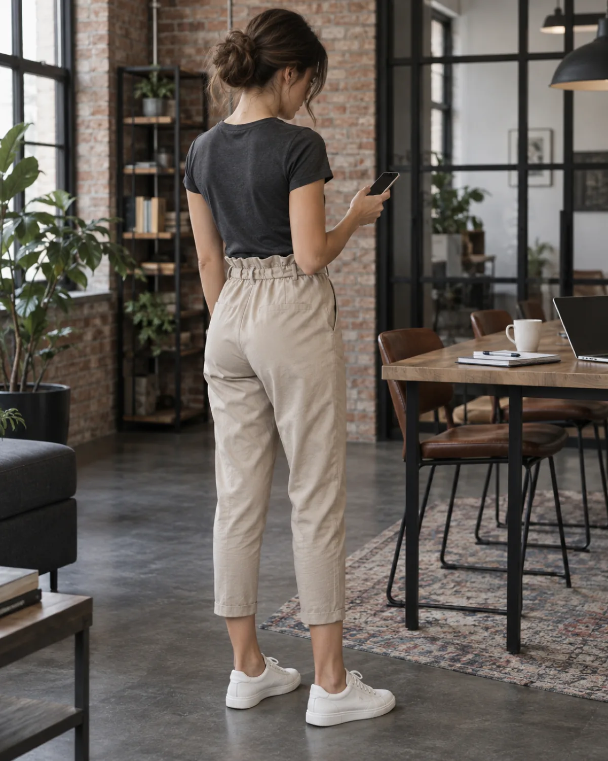 A petite woman seen from behind in a co-working space, looking at her phone, wearing stone paperbag-waist cropped trousers with a gathered waist, a charcoal fitted tee tucked in and white leather sneakers