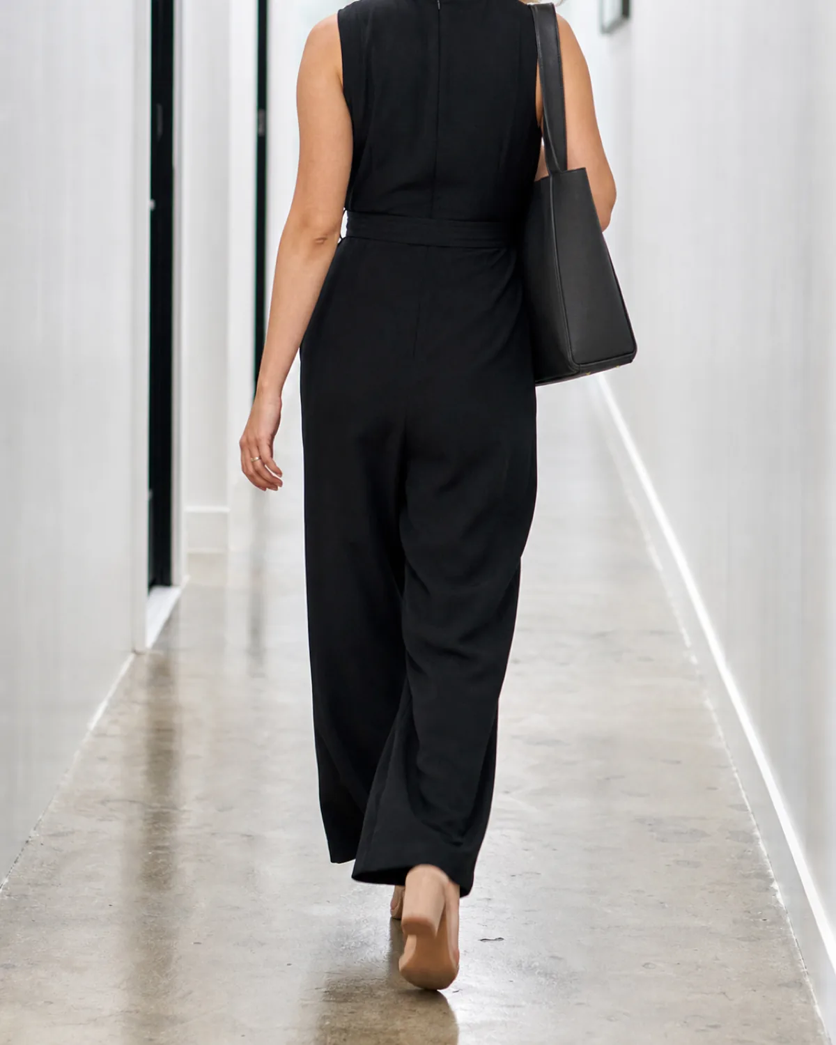 A woman walking away down a bright white hallway wearing a black wide-leg jumpsuit with a self-fabric belt and nude block heels, carrying a structured dark bag