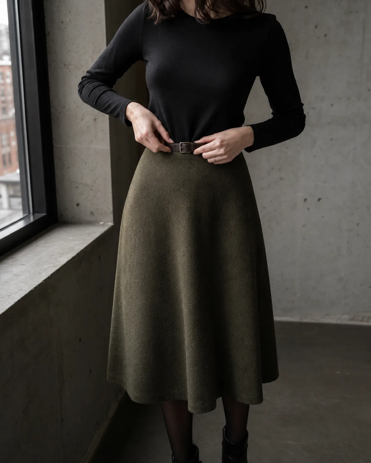 Mid-torso crop of a woman adjusting a dark leather belt at the waist of an olive A-line midi skirt with a fitted black crew-neck top and dark ankle boots