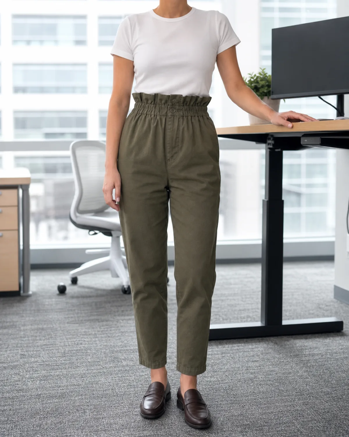 A woman standing in a bright office with one hand on a standing desk wearing olive paper-bag waist trousers with a fitted white crew-neck top and dark brown loafers