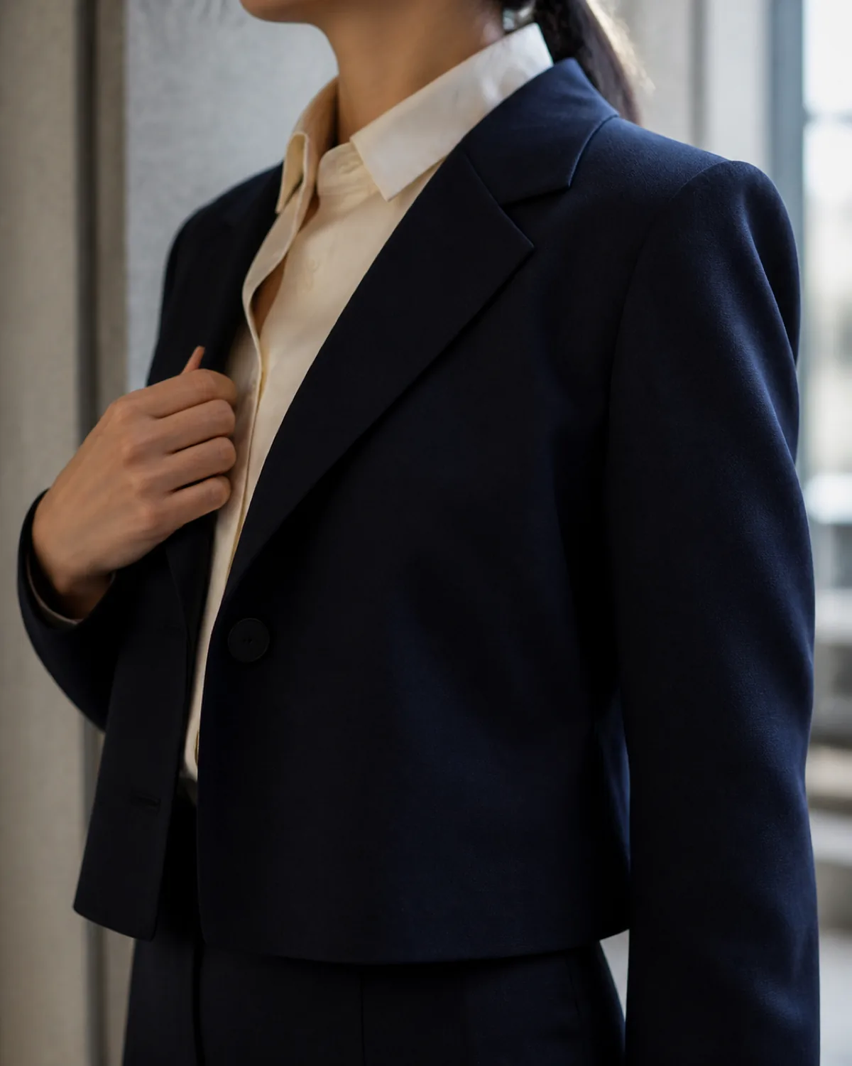 Close crop on the shoulder, collar and lapel of a navy short blazer over a cream silk blouse, with one hand adjusting the lapel
