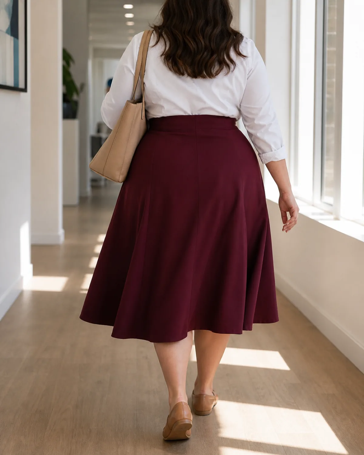 A plus-size woman walking away from the camera down a sunlit office hallway wearing a burgundy high-waisted A-line midi skirt, a white button-down shirt tucked in, and tan leather loafers, with a tan structured tote on her left shoulder