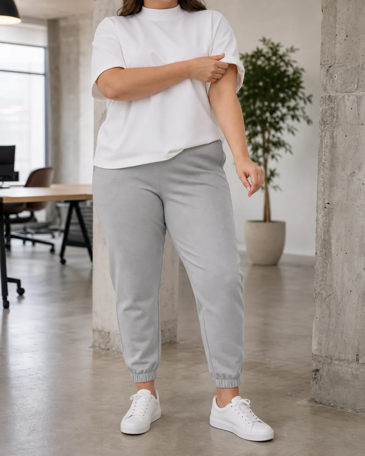 A plus-size woman in a bright creative office space with exposed concrete, wearing light grey tapered jogger trousers in ponte fabric, a white structured oversized tee, and white leather sneakers, her hands adjusting the rolled cuff of the right sleeve, head cropped at the chin
