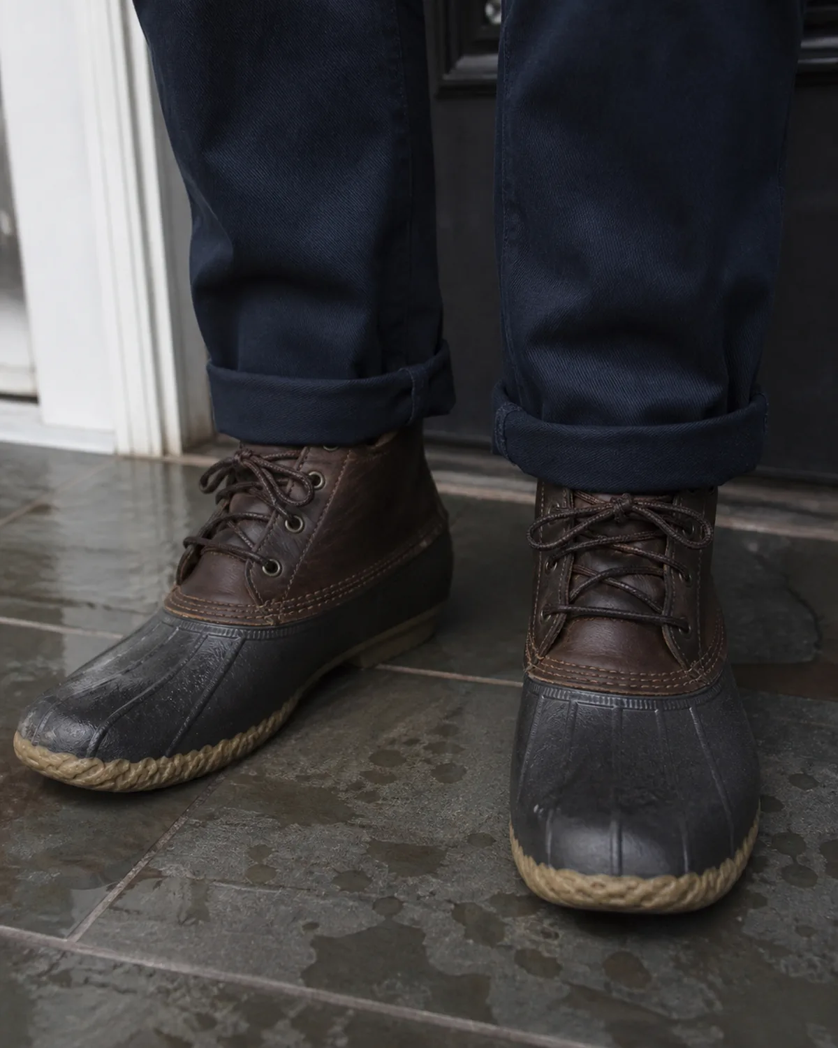 Close crop on the hem and shoes showing dark navy chinos with a single cuff above the ankle and dark brown duck boots with a rubber lower and leather upper standing on wet dark tile floor with a few small puddles