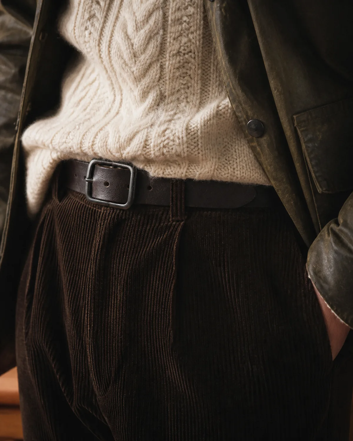 Close crop on the waist showing a cream cable-knit sweater partially tucked into dark brown corduroy trousers with a plain dark leather belt, the edge of an olive-brown waxed cotton jacket visible at the frame edges, visible corduroy wales and cable-knit texture in warm interior light