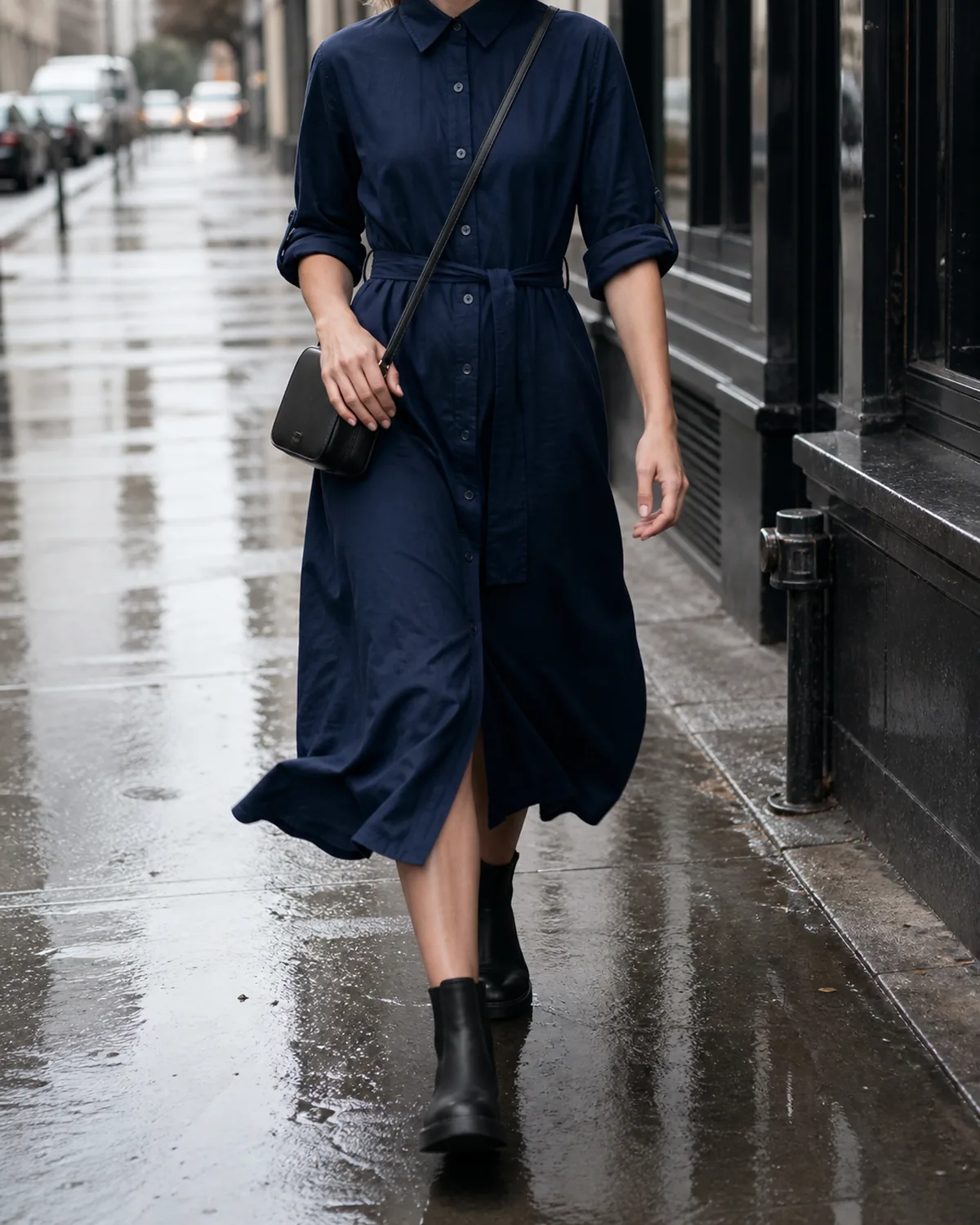 A woman walking mid-stride on a wet sidewalk in a navy cotton midi shirtdress with rolled sleeves and a self-fabric belt, wearing black waterproof Chelsea ankle boots and carrying a small black crossbody bag