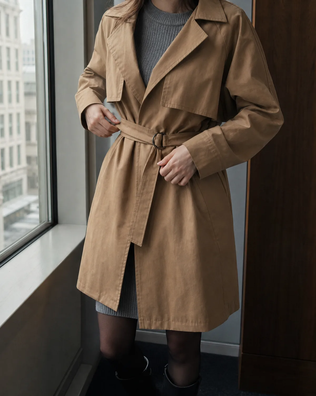 Mid-torso view of a woman adjusting the belt on a camel-coloured waterproof belted trench-style coat over a grey ribbed knit sweater dress, the dress visible at the neckline and below the coat hem, with dark tights and ankle boots at the bottom of the frame