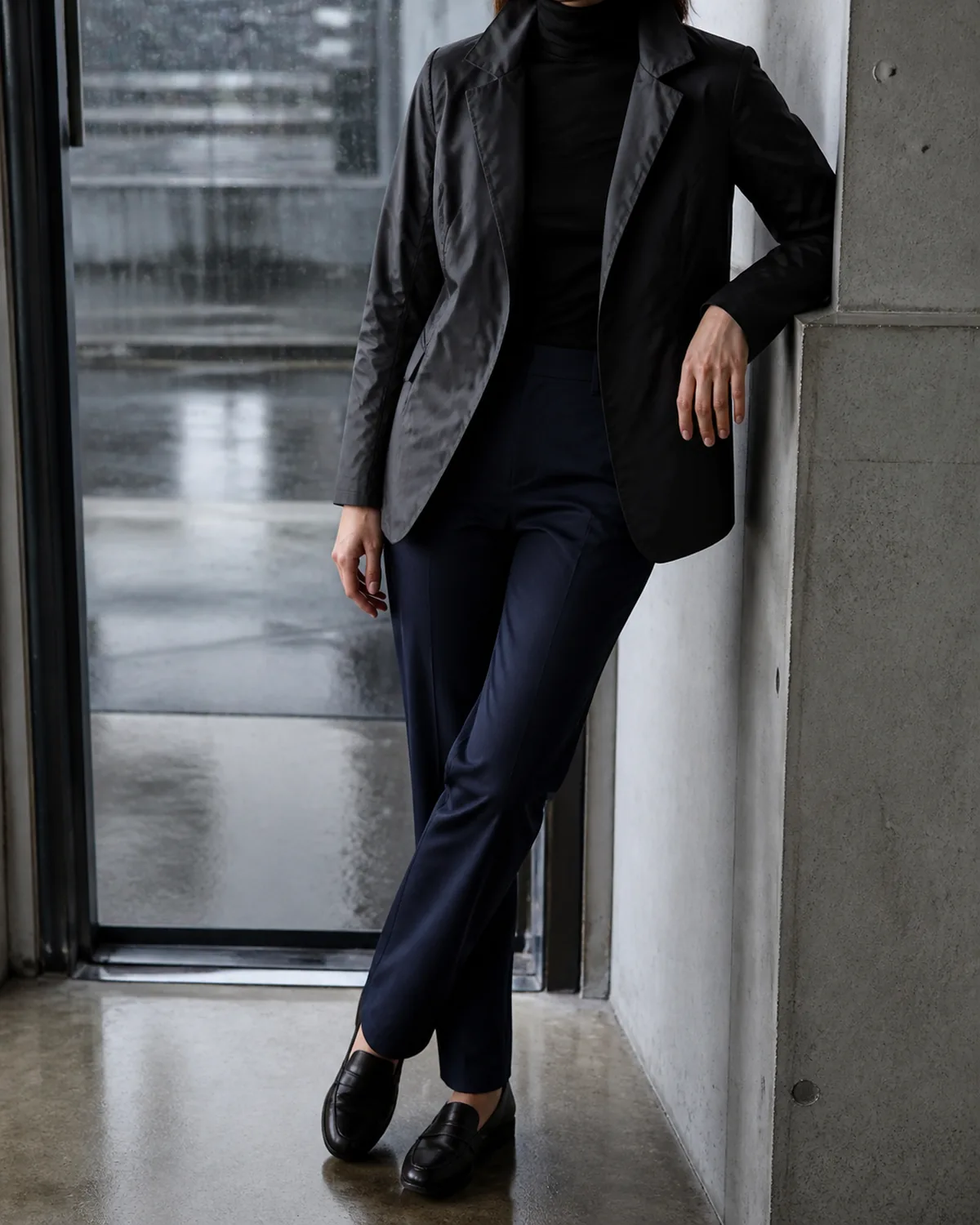 A woman leaning one shoulder against a concrete column in an office doorway, wearing a charcoal water-resistant blazer with a slightly shiny surface over a black fitted turtleneck and dark navy straight-leg trousers with black leather loafers, rain visible through the glass behind