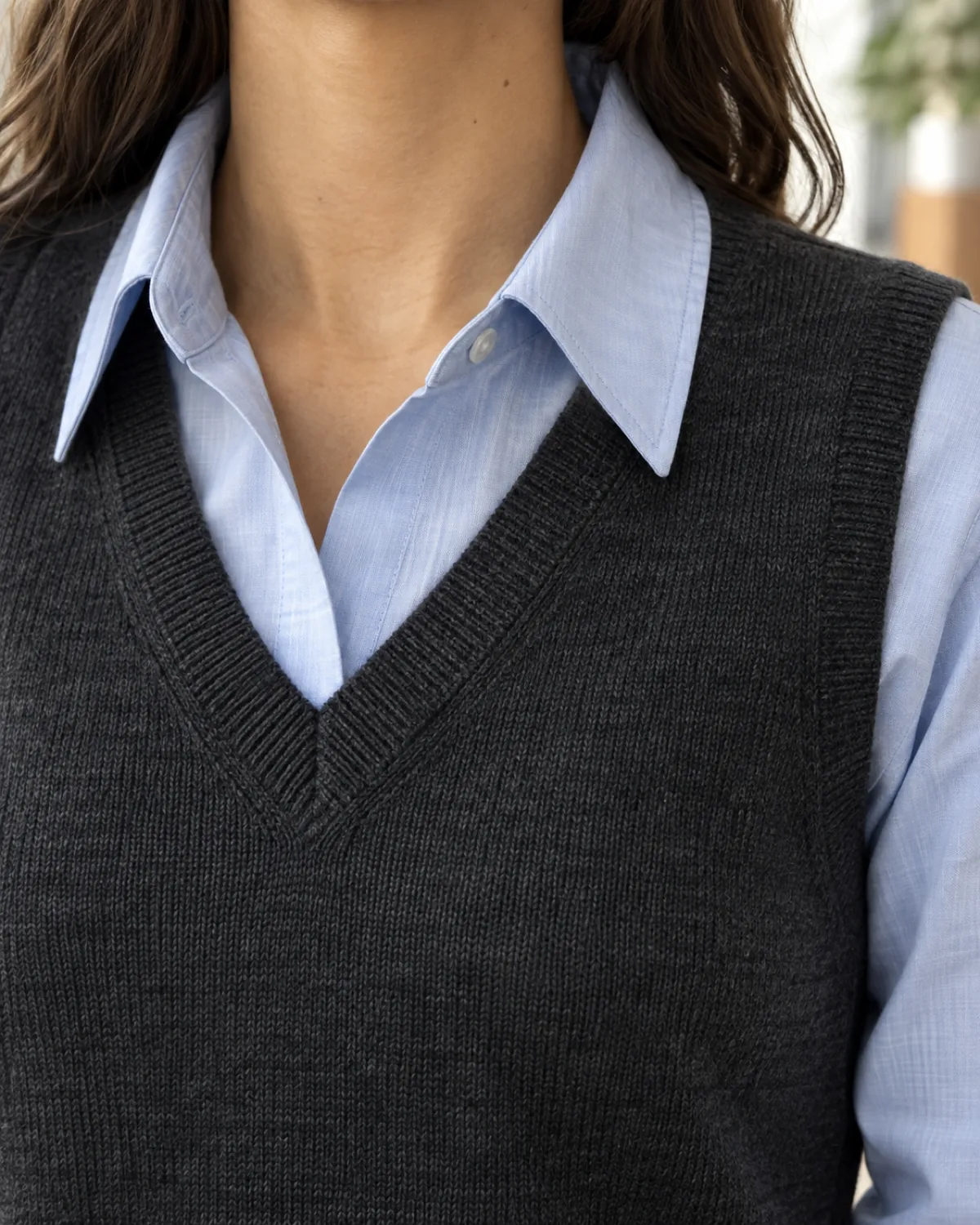 Close crop on the shoulder and collar area showing a charcoal V-neck fine-gauge knit vest over a light blue cotton button-down shirt with the collar visible above the vest neckline