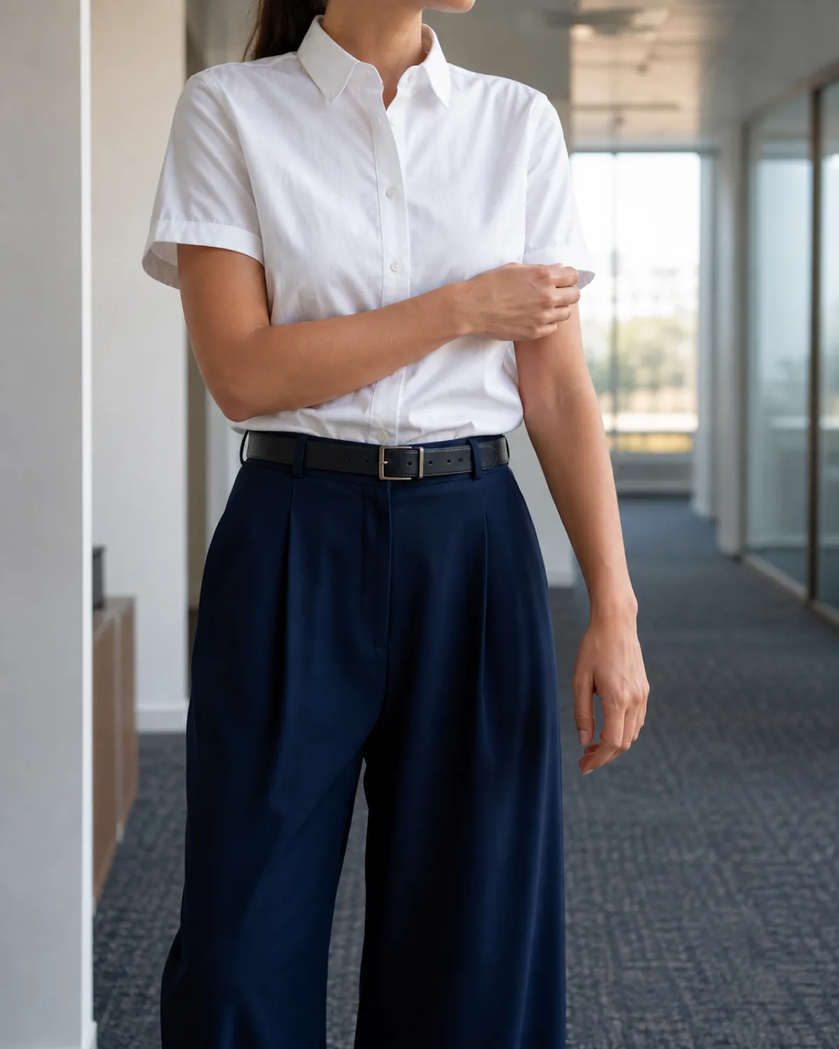 A woman in a white short-sleeve button-down shirt with a structured collar, tucked into navy wide-leg trousers with a dark belt, adjusting her cuff in an office corridor with neutral walls and cool window light