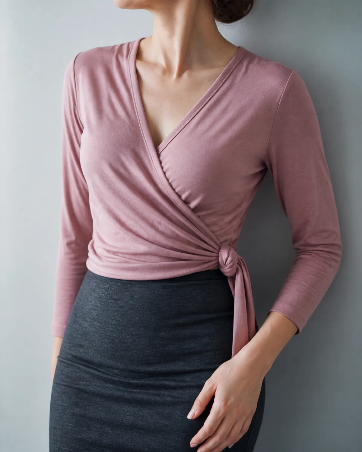 Close crop showing the crossover neckline and side tie of a dusty rose jersey wrap top over the waistband of a charcoal pencil skirt, against a plain neutral wall with soft cool light