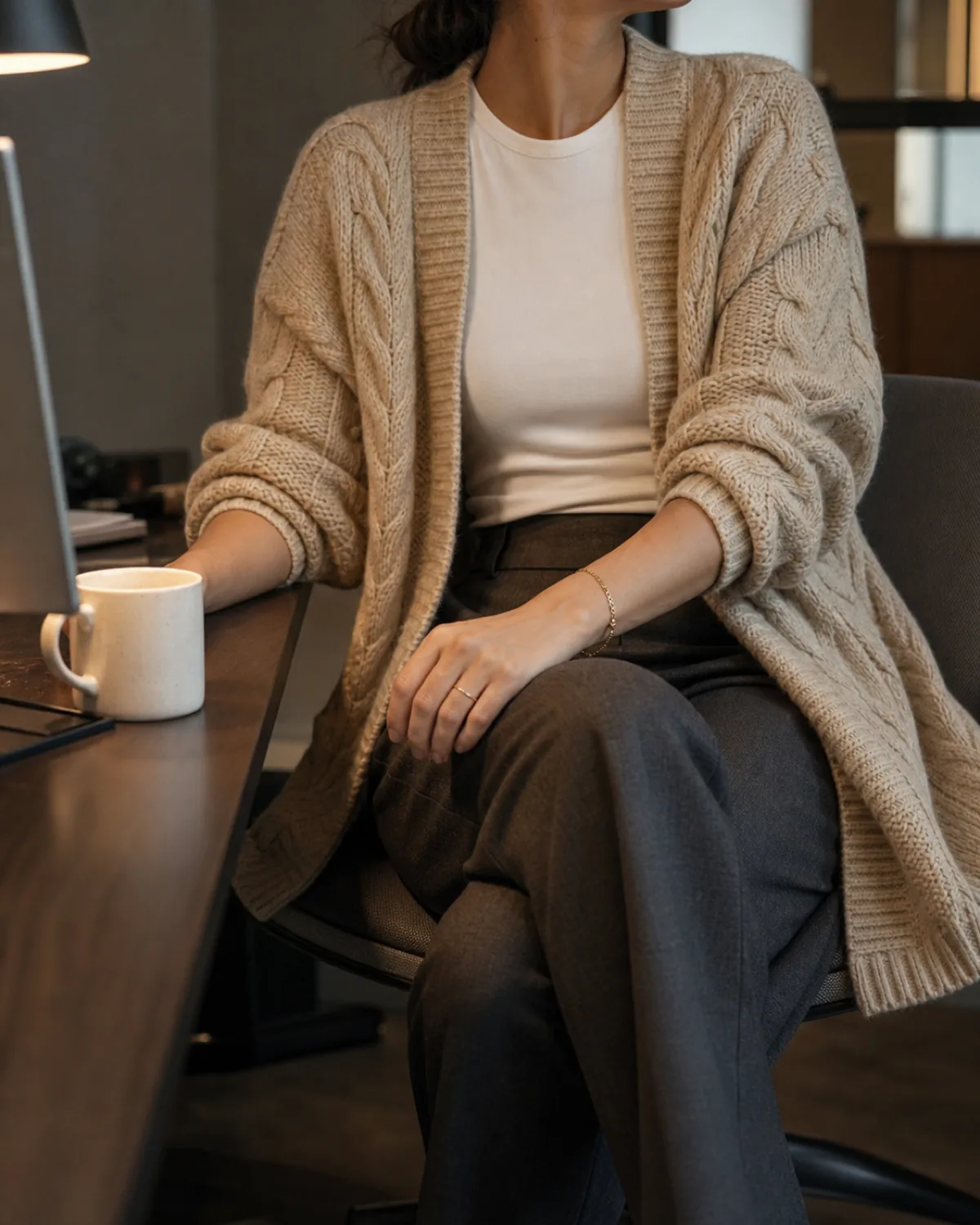 A woman seated at a modern office desk in an oatmeal chunky cable-knit cardigan over a white crewneck and dark grey trousers with a ceramic mug and monitor visible