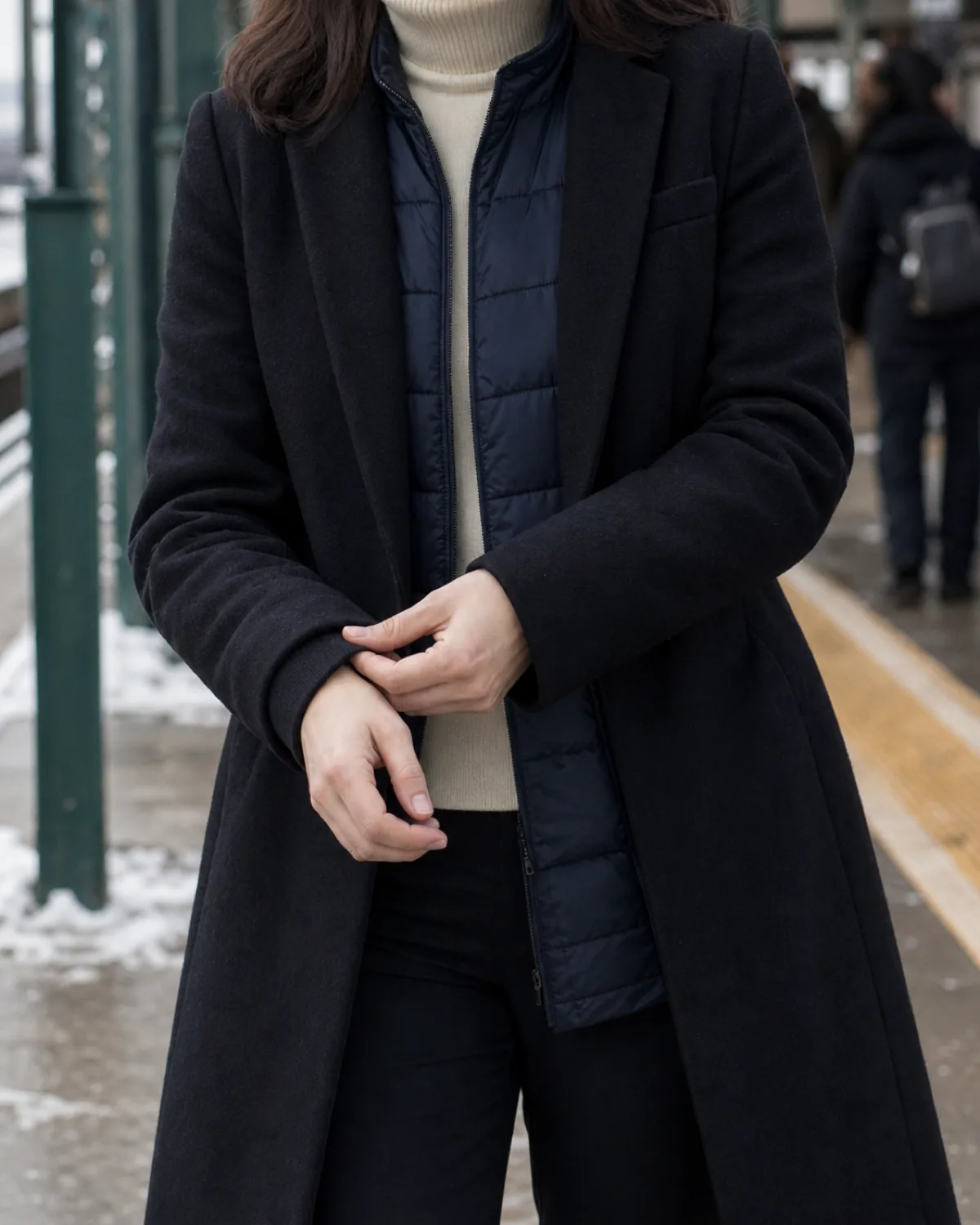 A woman at a winter transit platform adjusting her coat cuff, wearing a black wool topcoat open over a navy quilted puffer vest and cream turtleneck with dark trousers