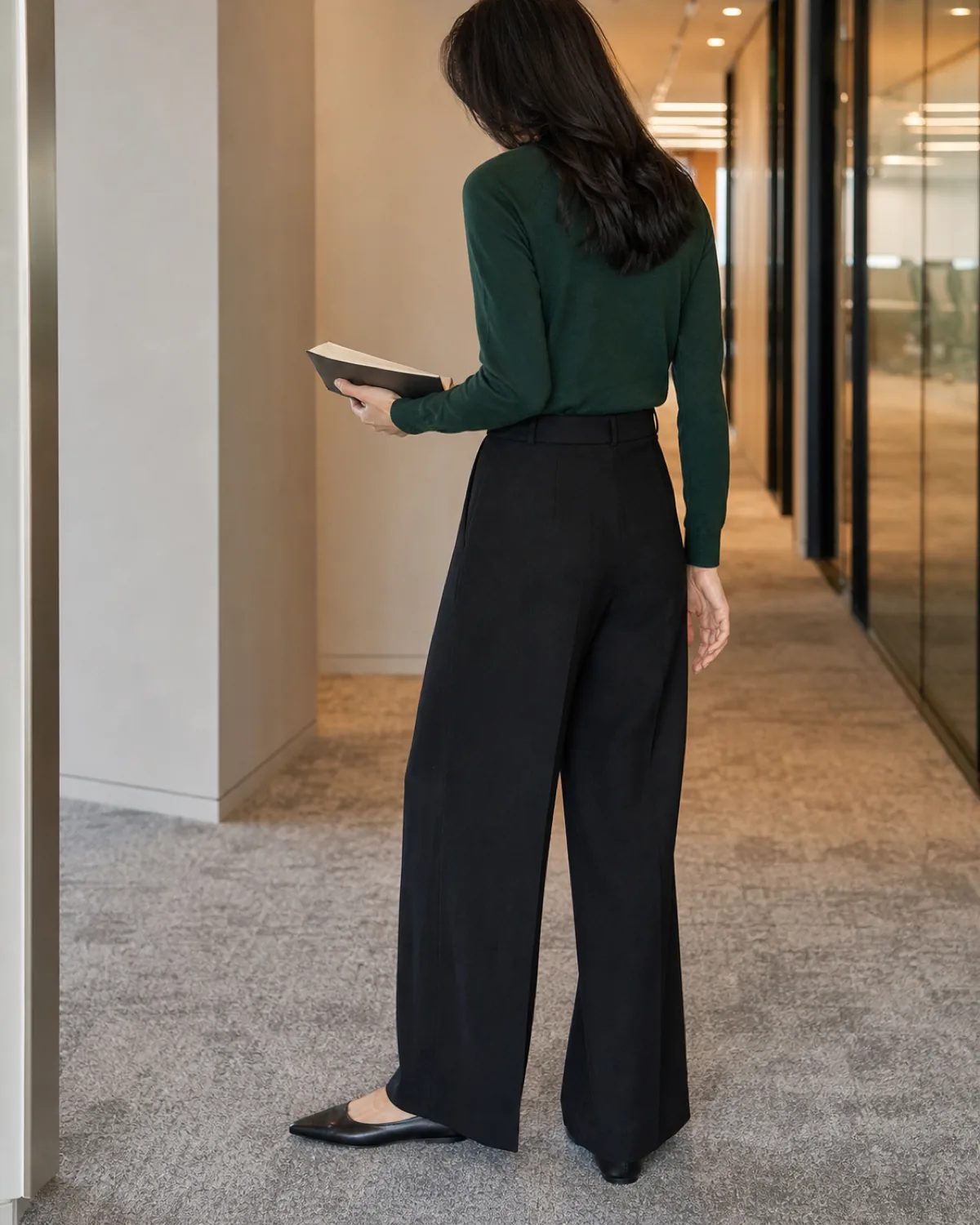 A woman seen from behind in a forest-green merino crewneck tucked into black wide-leg wool trousers with pointed-toe flats, holding a small notebook in an office corridor