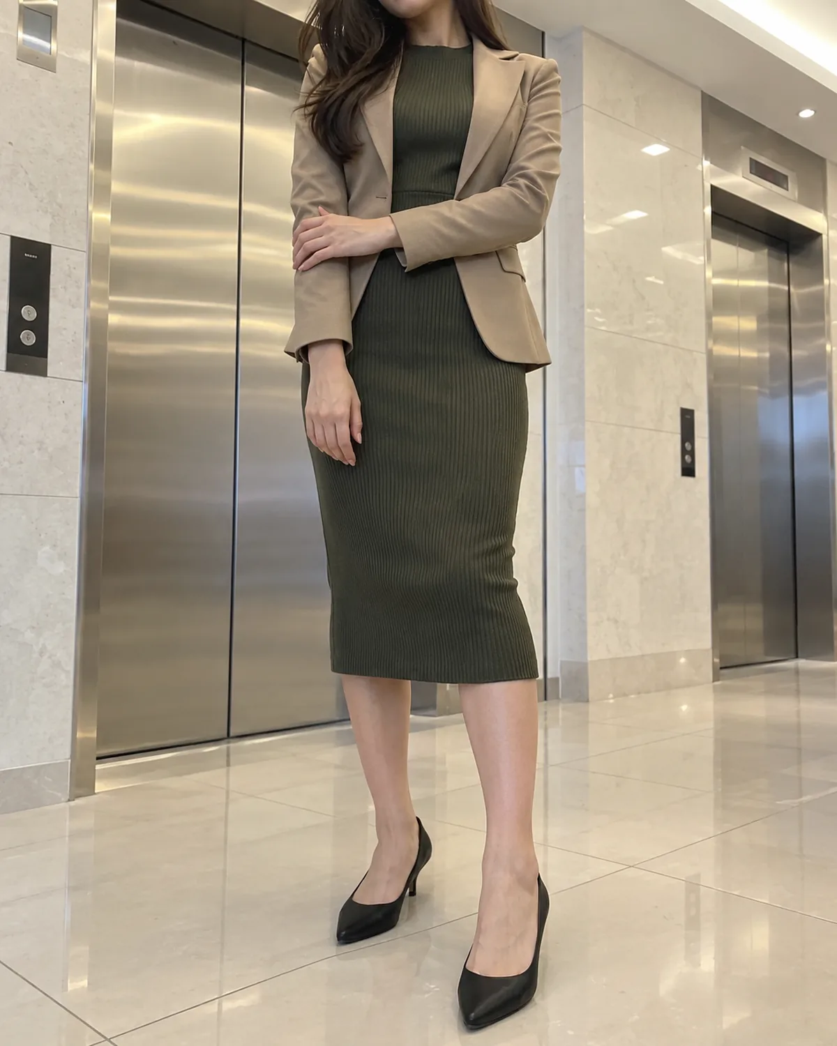 A petite woman standing in an elevator bay wearing a fitted dark-olive ribbed knit midi dress under a tan structured blazer with black pointed-toe low-heel pumps