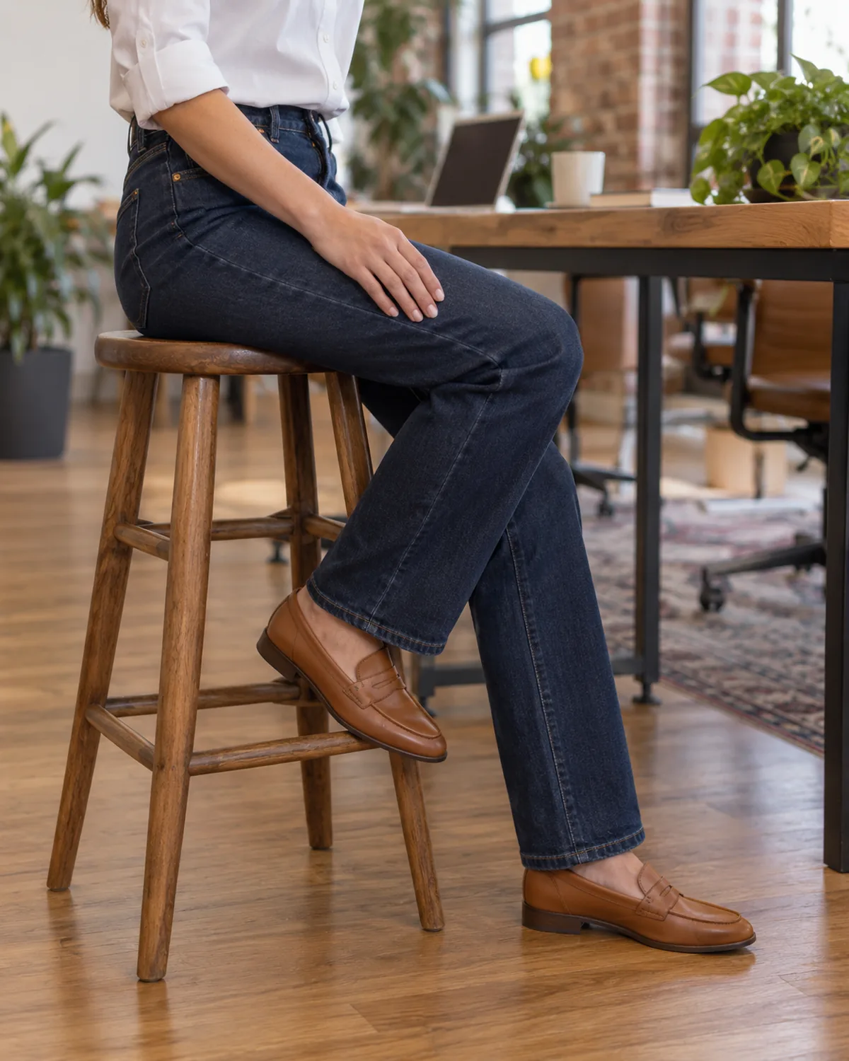 A petite woman seated on a wooden stool from the side in a casual office with exposed brick and plants, wearing dark-indigo straight-leg jeans with a white button-front blouse tucked in and tan leather penny loafers