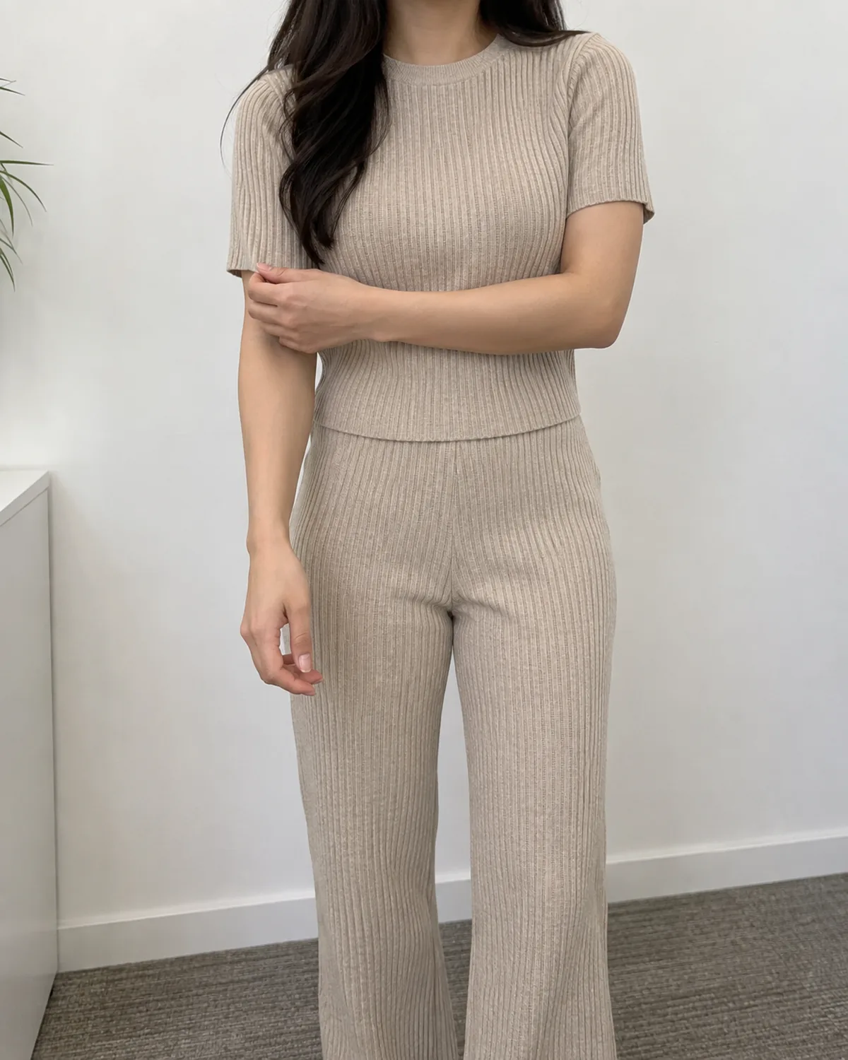 A petite woman adjusting her cuff while wearing a matching oatmeal ribbed knit crew-neck top and straight-leg knit trousers in an office setting