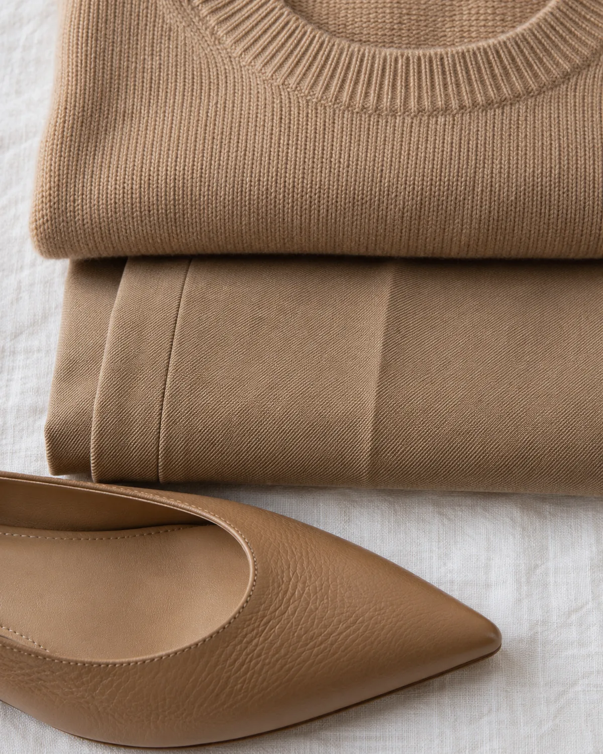 Stacked detail macro of three camel-coloured textures on a linen surface: the ribbed neckline of a folded crew-neck knit, a pressed trouser crease, and the toe of a leather pointed flat