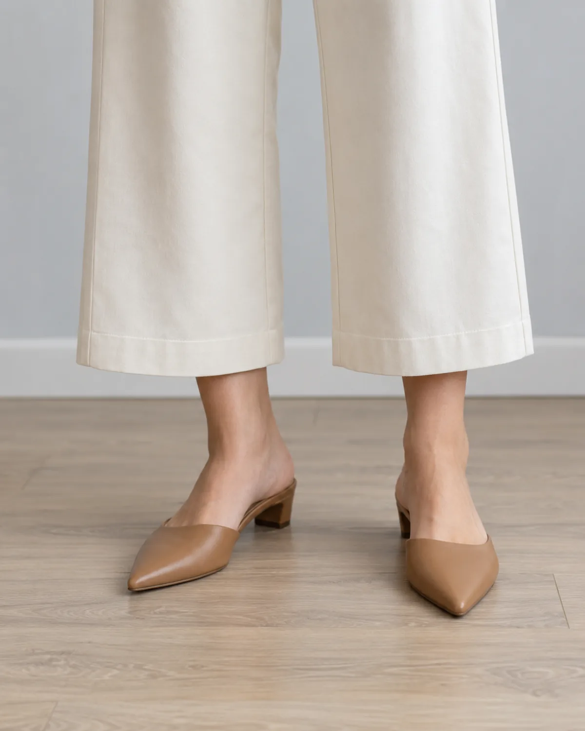 Close crop of cream wide-leg cropped trousers hemmed three inches above the ankle with tan leather pointed-toe mules on a light-wood office floor